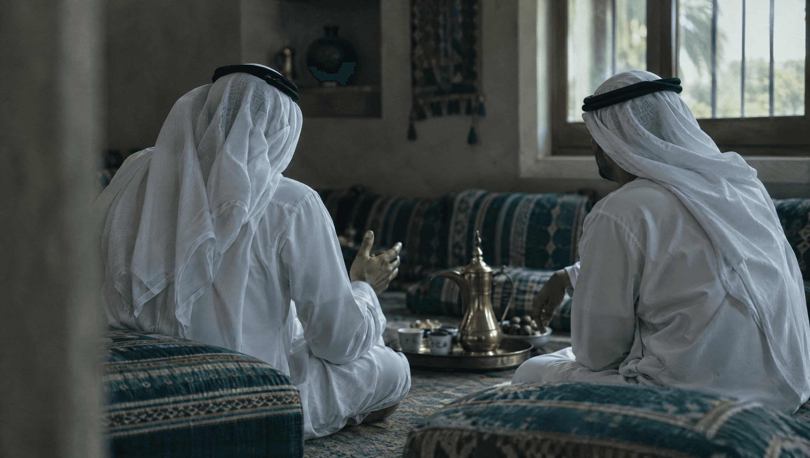 Two men in white kandura talking in a majlis, seen from behind, with a brass dallah coffee pot on a tray beside them.
